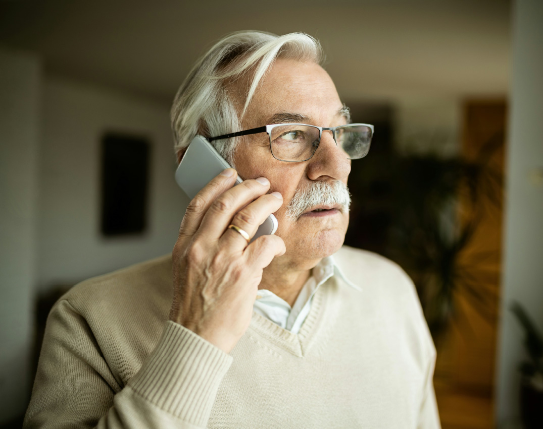 Ein älterer Mann mit nachdenklichem Gesichtsausdruck telefoniert mit einem Handy, scheinbar in seiner Wohnung.
