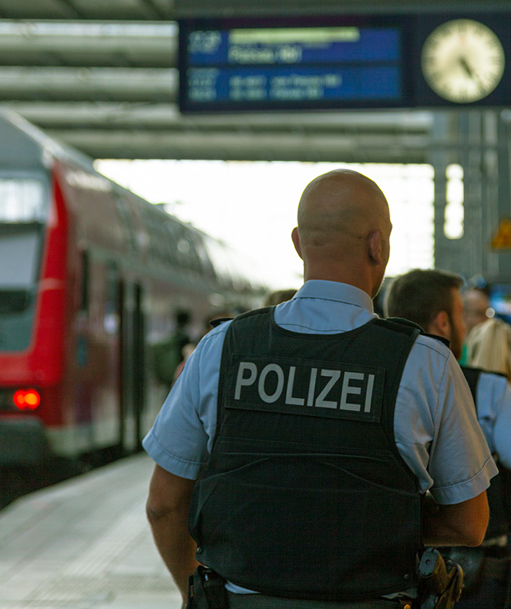 Mehrere Polizisten mit Schutzwesten stehen auf einem Bahnhofsgleis, im Hintergrund ein Zug und eine Anzeigetafel mit Uhr.
